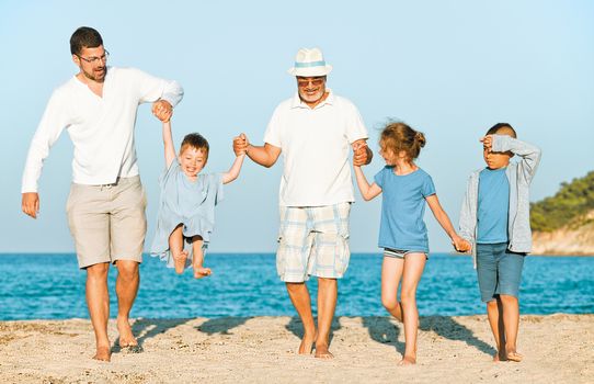 Family vacation on the beach. Group of five. Grandfather and father are carrying a toddler, two seven year young girl and a boy next to them, all holding hands.