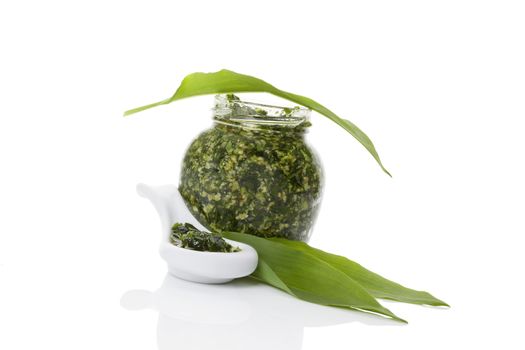 Garlic pesto in glass jar and white ceramic spoon with fresh wild garlic leaves isolated on white background. Modern minimal image style. Culinary healthy eating, spring detox. 