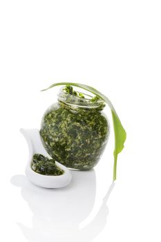 Garlic pesto in glass jar and white ceramic spoon with fresh wild garlic leaves isolated on white background. Modern minimal image style. Culinary healthy eating, spring detox. 