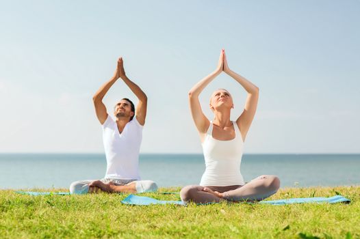 fitness, sport, friendship and lifestyle concept - smiling couple making yoga exercises sitting on mats outdoors