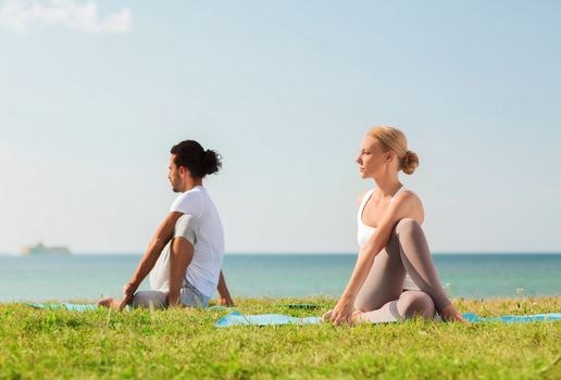 fitness, sport, friendship and lifestyle concept - smiling couple making yoga exercises sitting on mats outdoors