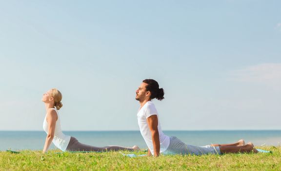 fitness, sport, friendship and lifestyle concept - smiling couple making yoga exercises lying on mats outdoors