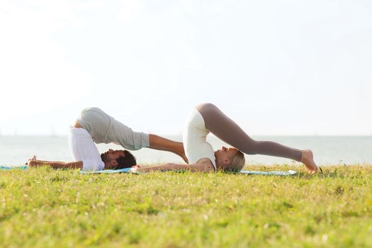 fitness, sport, friendship and lifestyle concept - smiling couple making yoga exercises on mats outdoors