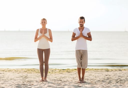 fitness, sport, friendship and lifestyle concept - smiling couple making yoga exercises on sand outdoors