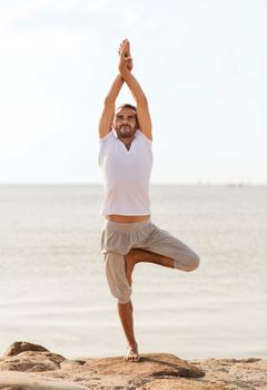 fitness, sport, people and lifestyle concept - smiling man making yoga exercises on beach