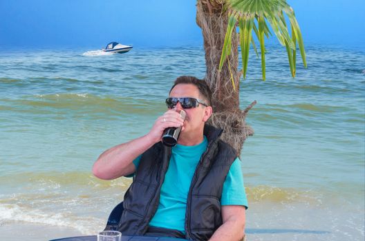 A man wearing sunglasses sitting in front of a palm tree. In the background the sea and a motorboat.