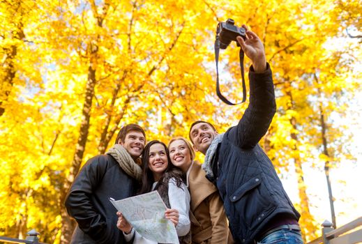 travel, people, tourism, technology and friendship concept - group of smiling friends with map and camera making self portrait in city park
