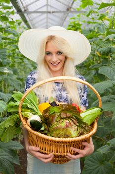 Attractive blond woman wearing a trendy straw sunhat standing holding a basket of fresh farm vegetables in a hot house while looking at the camera with a lovely warm smile