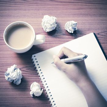 woman hand writing with pen on notebook.there are crumpled paper and coffee cup on wood table background vintage style