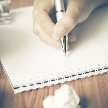 woman hand writing with pen on notebook.there are crumpled paper and coffee cup on wood table background vintage style