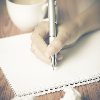 woman hand writing with pen on notebook.there are crumpled paper and coffee cup on wood table background vintage style