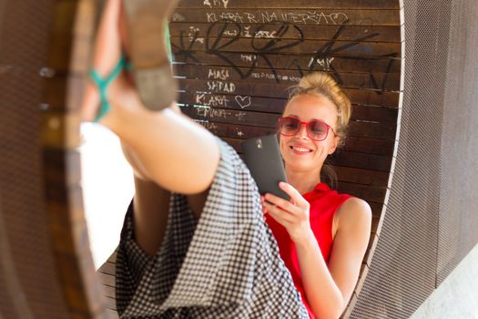 Casually dressed young cheerful lady wearing red sunglasses playing carelessly with her android smarthphone browsing trough social networks relaxing on  contemporary circular bench in city park.