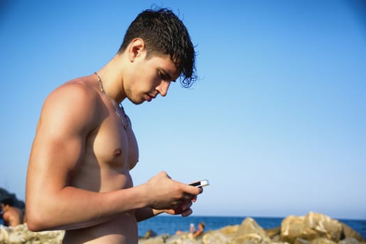 Shirtless Young Handsome Man Busy with his Mobile Phone While Standing at the Beach Boulders next to Sea or Ocean