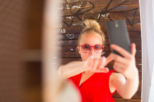 Casually dressed young cheerful lady wearing red sunglasses taking carefree selfie portrait outdoors with her android smarthphone to post it on social networks.