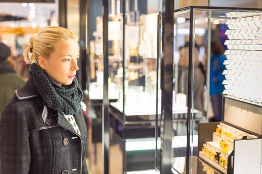 Beautiful blonde lady standing in front of showcase in beauty store, admiring new perfume collection.