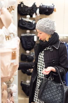 Woman shopping lingerie . Shopper looking and choosing clothing indoors in store. Beautiful blonde caucasian female model wearing winter coat and fashionable knitted cap.