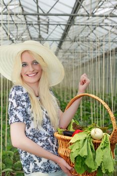 Pretty Blond Woman Wearing Straw Hat, Carrying Basket of Fresh Veggies on her Arm Inside the Farm and Looking Into the Distance.