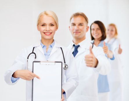 healthcare and medicine concept - smiling female doctor pointing finger to clipboard and blank page with team on the back