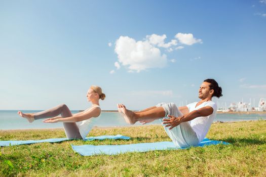 fitness, sport, friendship and lifestyle concept - smiling couple making yoga exercises sitting on mats outdoors