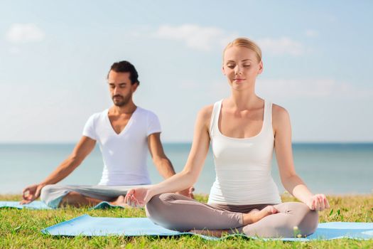 fitness, sport, friendship and lifestyle concept - smiling couple making yoga exercises sitting on mats outdoors