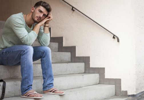 Casual  handsome young man with a beard sitting on cement steps with his head resting on his hand and a serious dejected expression