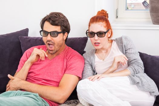 Young Couple Wearing 3D Glasses, Sitting on the Couch in the Living Room, Watching a Movie and Showing Shocked Faces.