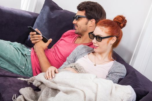 Young Couple Wearing 3D Glasses, Sitting on the Couch in the Living Room, Watching a Movie and Showing Shocked Faces.