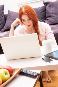 Pretty Young Woman Using her Laptop Computer In the Living Room with Feet on the Table and Showing Serious Face.