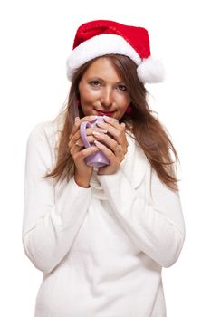 Cold attractive young woman with a cute smile in a festive red Santa hat sipping a hot mug of coffee that she is cradling in her hands to warm up in the winter weather, on white