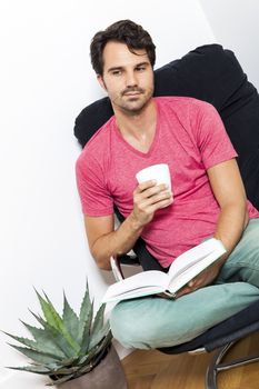 Young Man in Casual Clothing Sitting on Black Chair While Reading a Book and Holding a Glass of Drink.
