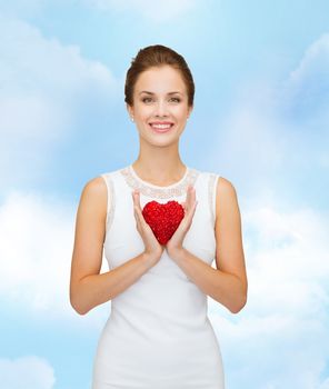 happiness, health, charity and love concept - smiling woman in white dress with red heart over blue cloudy sky background