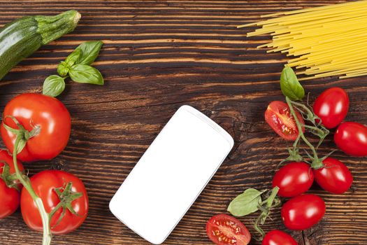 Fresh vegetables and pasta on wooden table with smartphone and empty white screen. 