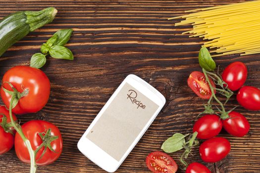 Fresh vegetables and pasta on wooden table with smartphone and recipe on screen. 