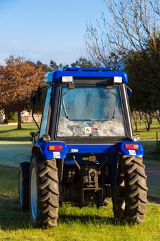 Rear view of a small blue tractor parked on grass with trees in the background.