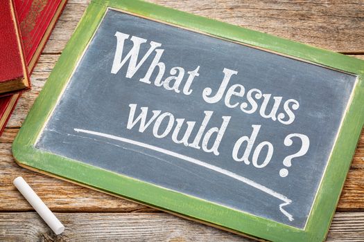 What Jesus would? An inspirational question on a slate blackboard with a white chalk and a stack of books against rustic wooden table