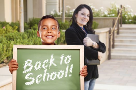 Happy Hispanic Boy Holding Back to School Chalk Board Outside on School Campus as Teacher Looks On.