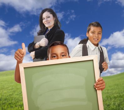 Young Hispanic Boys with Blank Chalk Board and Teacher Behind on Grass Field.