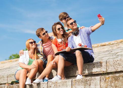 friendship, leisure, summer, technology and people concept - group of smiling friends with skateboard and smartphone making selfie outdoors