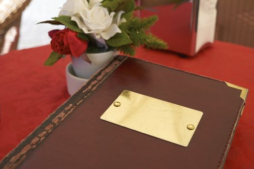 Detail of leather bound menu with blank name plate on red table cloth. There's a small flower vase on the side with red and white roses.