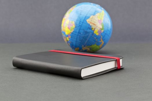 Journal with red elastic placed with globe on sophisticated gray background shows theme of travel, world wide exploration, adventure, and memories. Black book reflects opportunities for business and data records. 