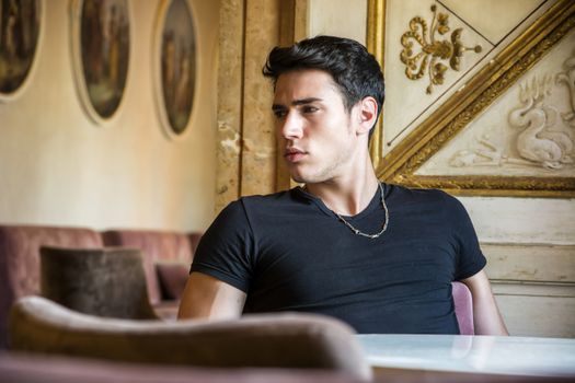 Portrait of Serious Attractive Young Man Sitting at Table Leaning on it and Looking Away, in Room Decorated with Gold Framed Artwork