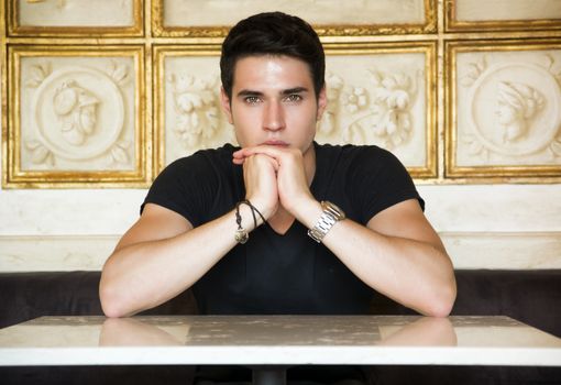 Portrait of Serious Attractive Young Man Sitting at Table Leaning on it and Staring at Camera in Room Decorated with Gold Framed Artwork