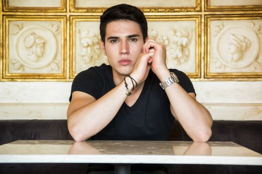 Portrait of Serious Attractive Young Man Sitting at Table Leaning on it and Staring at Camera in Room Decorated with Gold Framed Artwork