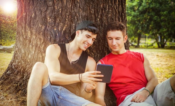 Two young male friends with casual trendy summer clothes leaning on an old tree trunk, while watching something funny on a tablet PC or ebook reader, in a warm day in the park