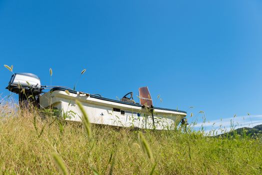An old abandoned boat surrounded by overgrown grass with a clear blue sky, small clouds and mountains in the distant background.