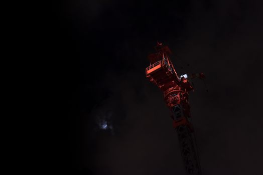 A large crane lit with a spotlight on a very dark night sky background next to a small moon behind clouds.