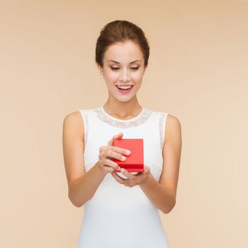 holidays, presents, wedding and happiness concept - smiling woman in white dress holding red gift box over beige background