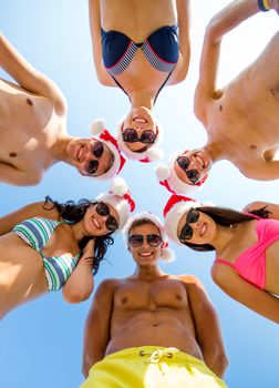 friendship, christmas, summer vacation, holidays and people concept - group of smiling friends wearing swimwear and santa helper hats standing in circle over blue sky