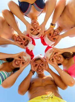 friendship, christmas, summer vacation, holidays and people concept - group of smiling friends wearing swimwear and santa helper hats standing and having fun in circle over blue sky