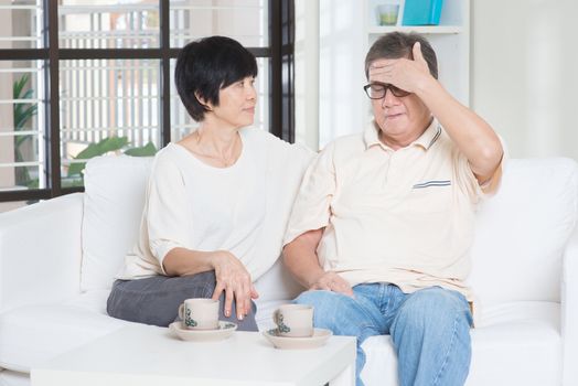 Portrait of mature Asian man having headache, sitting on sofa with wife at home, senior retiree indoors living lifestyle.
